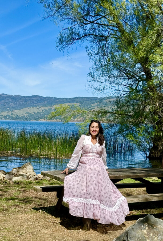"Trinity, tantric dominatrix, seated by a lake in a pink floral dress, receiving devotional gifts with grace.