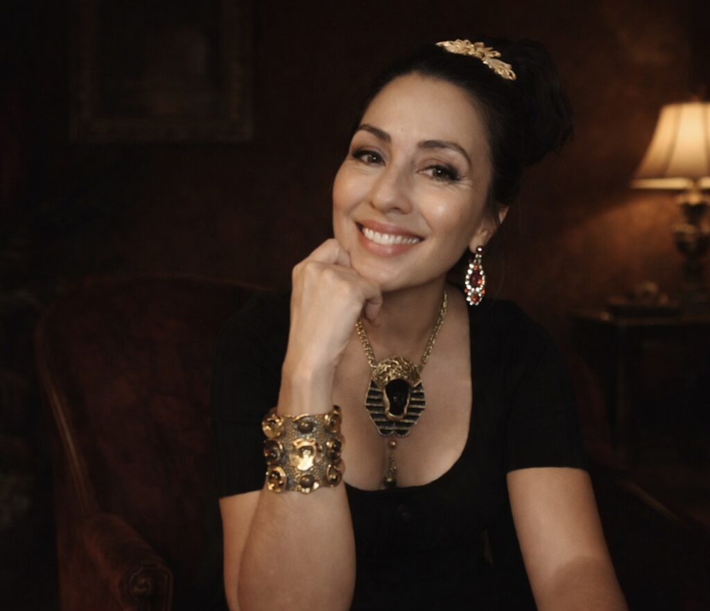 Trinity seated in gold jewelry and black dress, embodying sovereign feminine presence